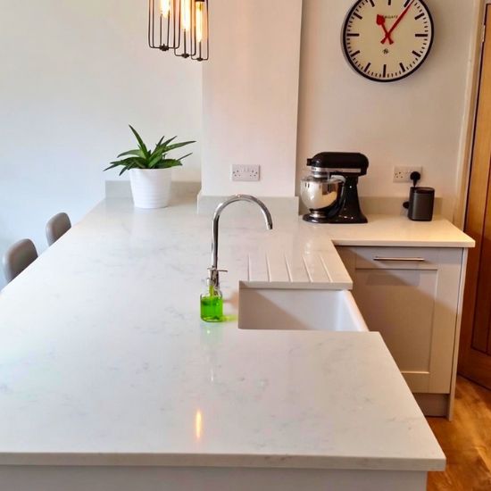 White Carrara Light Giant Quartz kitchen island worktop in bright white with subtle light grey veins, featuring undermount sink, chrome faucet, soap dispenser, potted plant, and stand mixer in a modern bright kitchen