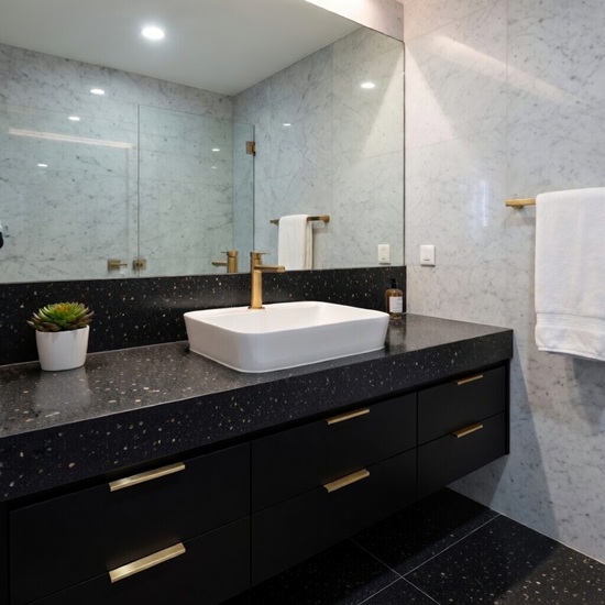 Quartzforms Terrazzo Doge quartz bathroom vanity top in deep black with white pink and copper specks, featuring white rectangular sink, gold faucet, black cabinet and marble walls