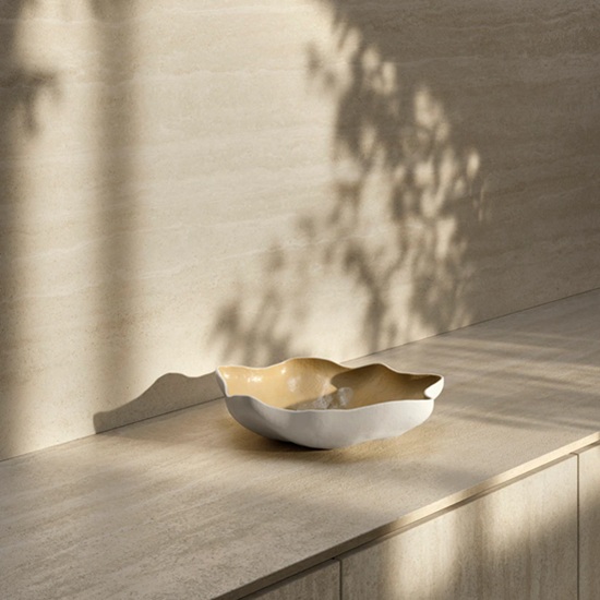 Sapienstone Travessa worktop in soft beige travertine-effect with subtle linear veining, featuring a sculptural bowl under dramatic natural light and shadows